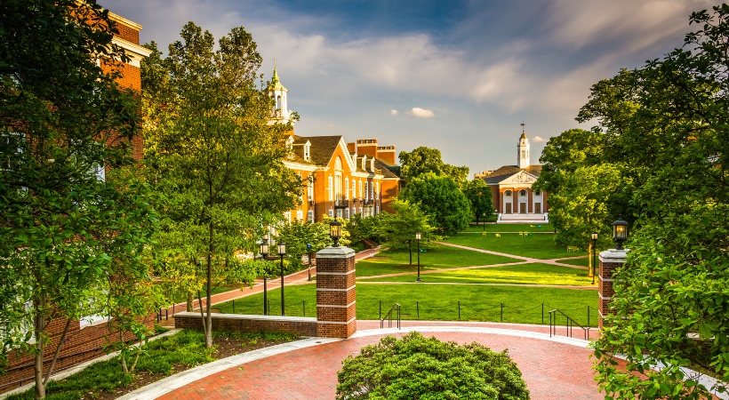 A tree-lined campus or park setting in spring showing mature trees with full green foliage surrounding brick buildings and manicured lawns.